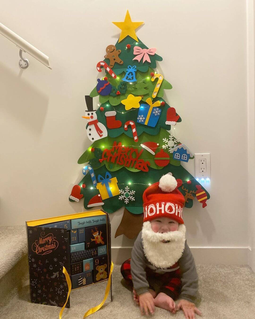 Bebê branco com barba e gorro do papai noel sentado em frente da árvore de natal em feltro